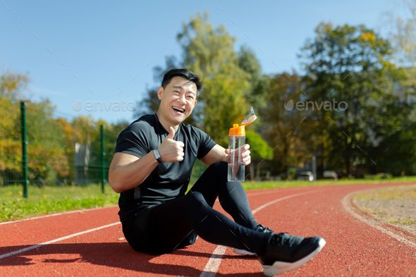 An asian sportsman, athlete, runner sitting on treadmill and resting after jogging. He holds a - Stock Photo - Images