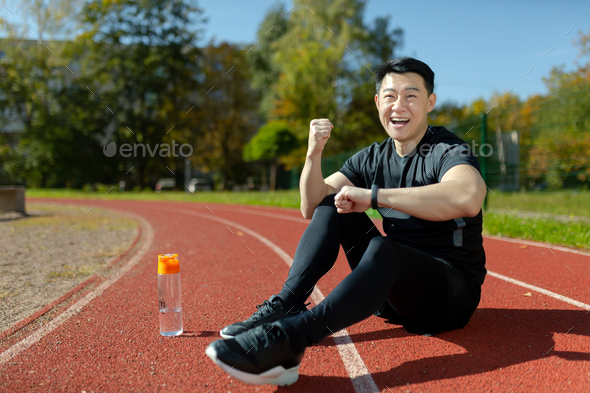 A young Asian man, an athlete sitting on a stadium treadmill. He looks at the smartwatch, fitness - Stock Photo - Images