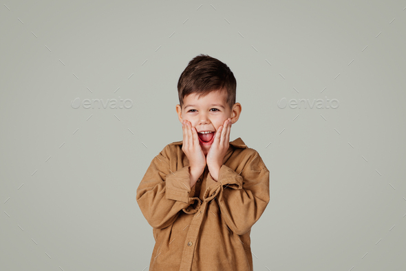 Cheerful shocked small european boy with open mouth holding hands to face - Stock Photo - Images