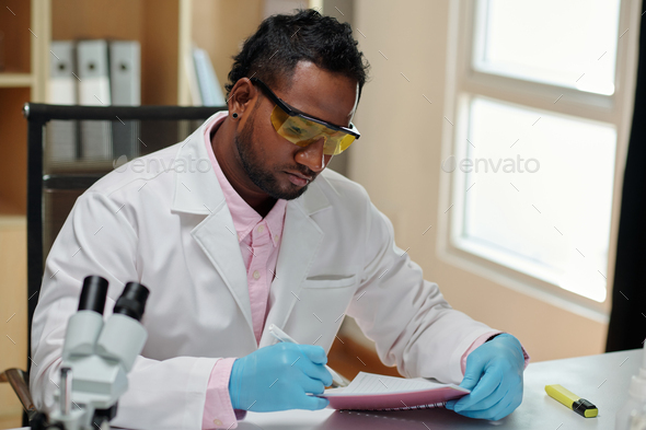 Young man in lab coat and gloves making notes in copybook - Stock Photo - Images