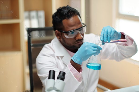 Young black man in workwear pouring liquid from flask into beaker - Stock Photo - Images