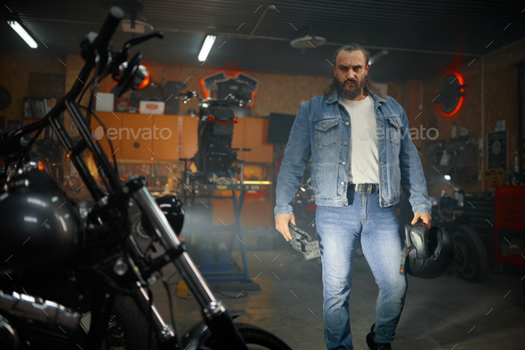 Portrait of bearded brutal biker looking motorcycle in garage workshop - Stock Photo - Images