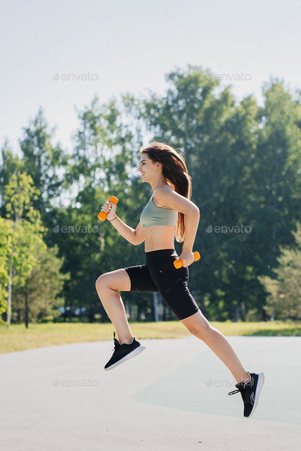 Vertical shot of brunette hispanic young woman jumps high holds dumbbells against blurrypark.