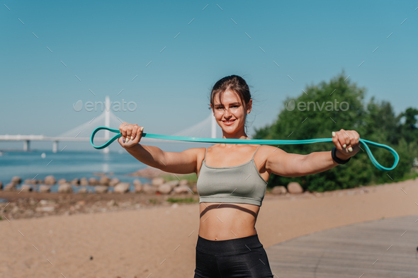 Fit brunette hispanic girl in sportswear stretching rubber tapes, doing workout at beach, bay