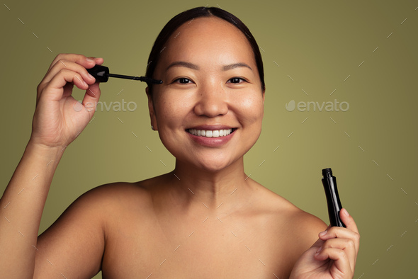 Joyful young Asian woman applying mascara in green studio - Stock Photo - Images
