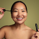 Joyful young Asian woman applying mascara in green studio - PhotoDune Item for Sale