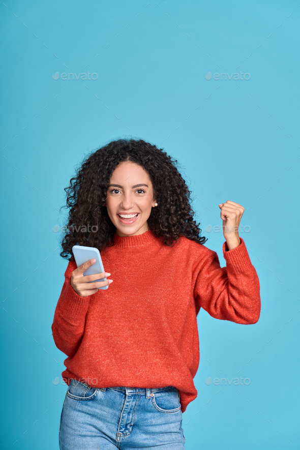 Young happy latin woman winner holding mobile cell phone isolated on blue. - Stock Photo - Images