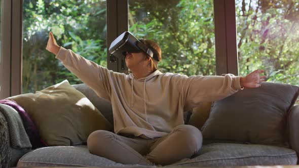 Asian girl smiling and gesturing while wearing vr headset sitting on the couch at home alt