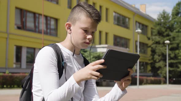 A Caucasian Teenage Boy Works on a Tablet with Earphones on  a School in the Background