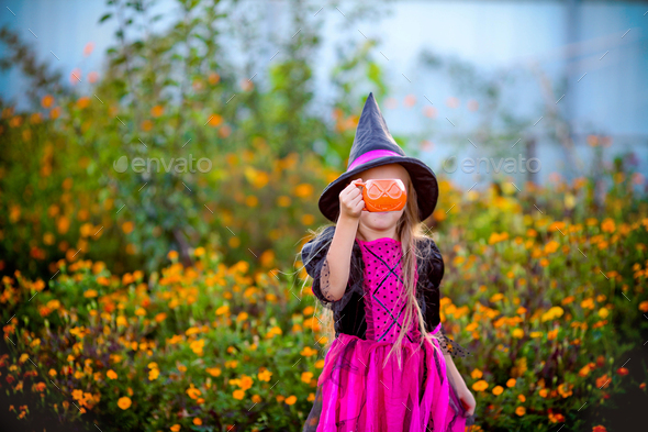 A little girl dressed as a witch stands in the street and hides her face with a pumpkin for sweets.