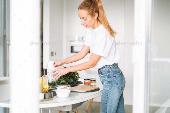 Young slim woman in white t-shirt and blue jeans cooking smoothie healthy food in kitchen at home - Stock Photo - Images