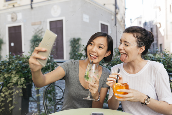Italy, Padua, two young women pulling funny faces while taking selfie at sidewalk cafe