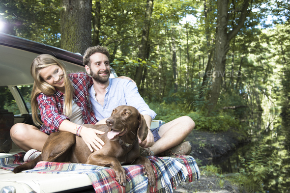 Smiling young couple with dog sitting in car at a brook in forest
