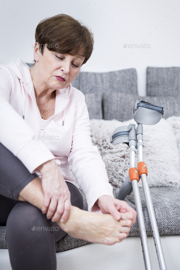 Senior woman with crutches sitting on couch, checking her ankle