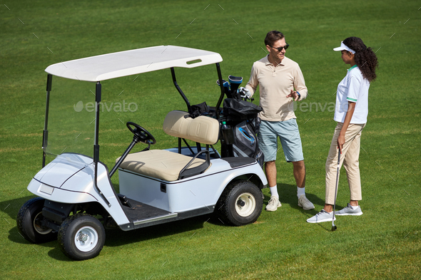 Elegant couple standing by golf cart on green field and chatting in sunlight - Stock Photo - Images