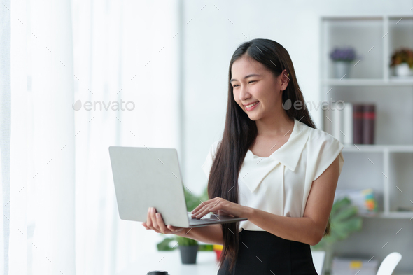 Beautiful Asian business woman standing holding a laptop in the office - Stock Photo - Images