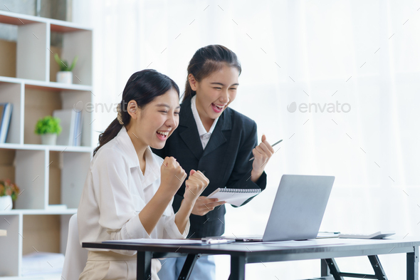 Two beautiful young Asian businesswomen raising their hands to express their joy at their satisfying - Stock Photo - Images