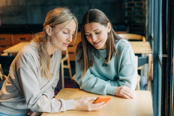 Two modern women surf the internet using a mobile phone while sitting in a coffee shop. - Stock Photo - Images