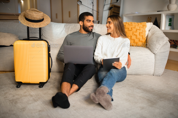 Smiling international millennial couple sit on floor with gadgets, suitcase and hat, choose country - Stock Photo - Images
