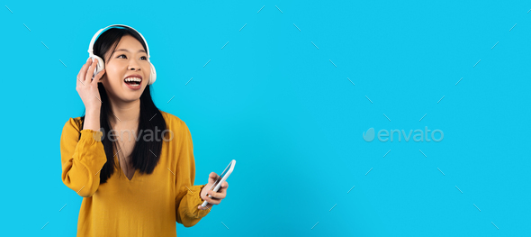 Joyful asian woman listening to music in headphones, using smartphone - Stock Photo - Images