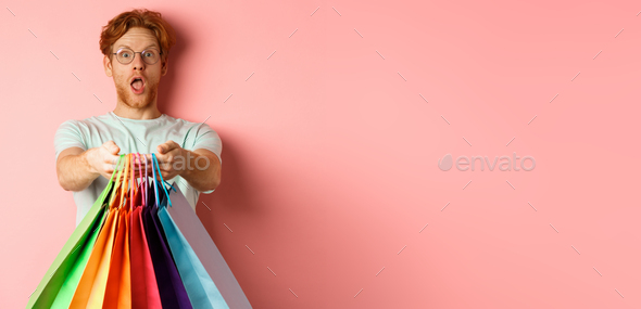Surprised redhead man stretch out hands with shopping bags, give you gifts, standing over pink