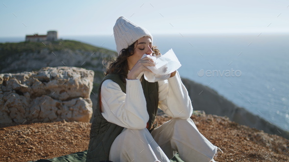 Girl hiker eating food at ocean cliff top. Serene travel breakfast at ocean view