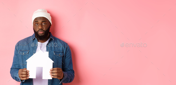 Real estate concept. Sad and tired Black man staring unamused at camera, holding paper house model