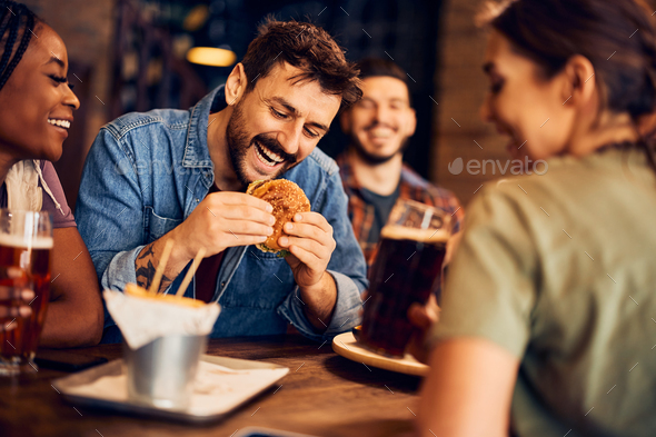 Happy man eating burger while gathering with friends in a bar. - Stock Photo - Images
