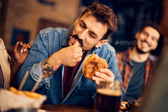 Hungry man eating burger while gathering with friends in a pub.