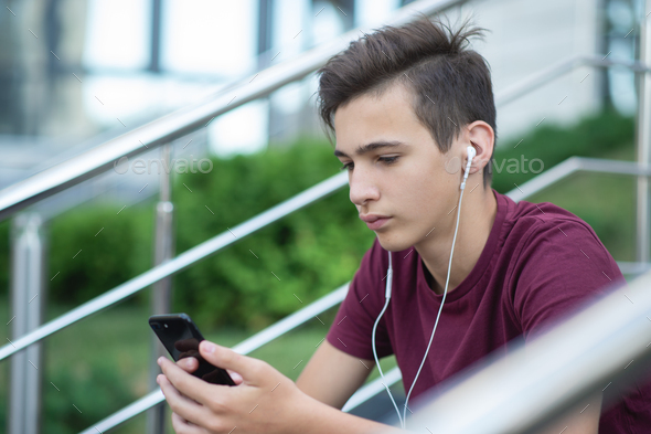 Teenage boy is using mobile phone, outdoors. Handsome young man with smartphone and earphones.  - Stock Photo - Images