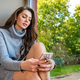 young sensual millennial long-haired brunette with smartphone sitting on the windowsill at home - PhotoDune Item for Sale
