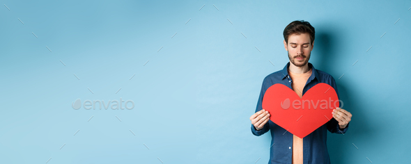 Lonely guy looking sad at valentines red heart with sad face, standing over blue background