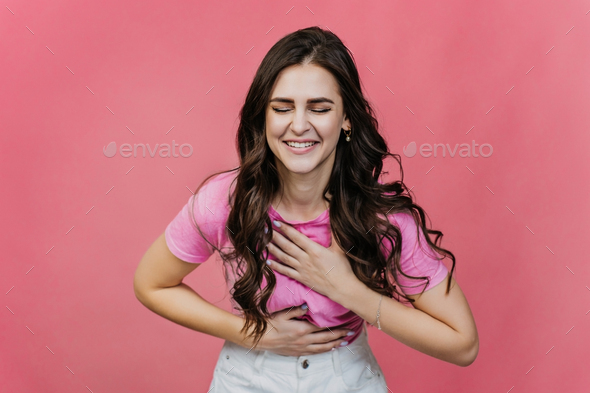 Fit hispanic young woman with loose wavy hair laughs eyes closed over pink studio backdrop.