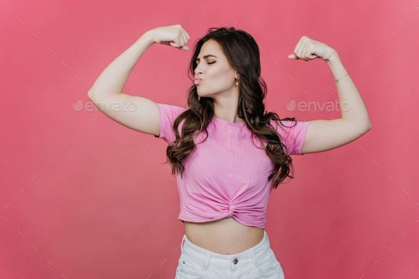 Fit hispanic young woman with loose wavy hair shows her biceps over pink studio backdrop. Strength