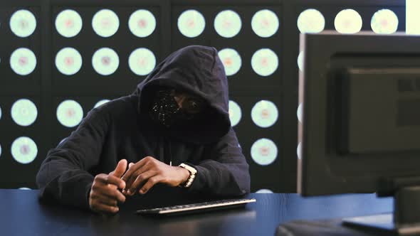 Africanamerican Male Hacker in Black Face Mask and Hoodie Puts Smartphone Away
