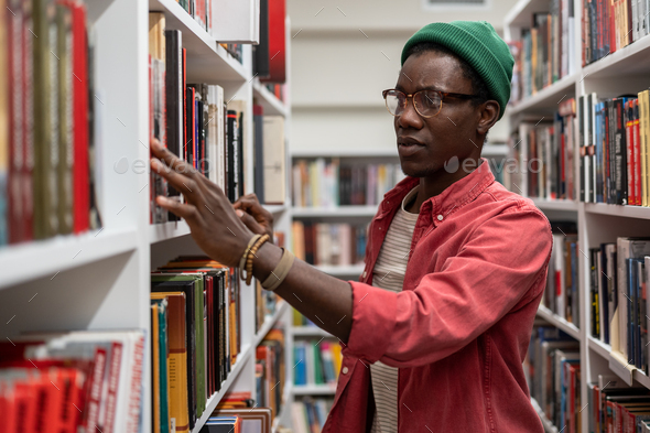 African guy University student standing between bookshelves in library - Stock Photo - Images