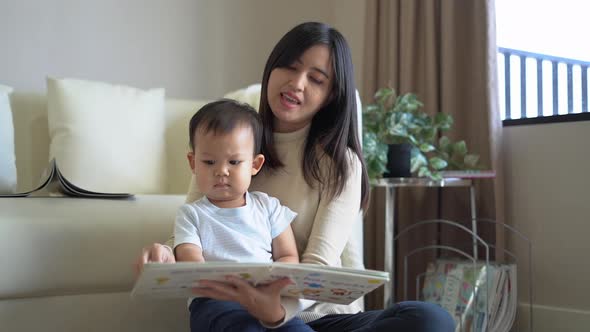 Young mother and little boy child reading book sitting on floor near sofa at home. Happy family.