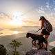A sporty girl with a backpack stands on the edge of a mountain with a Rottweiler dog - PhotoDune Item for Sale