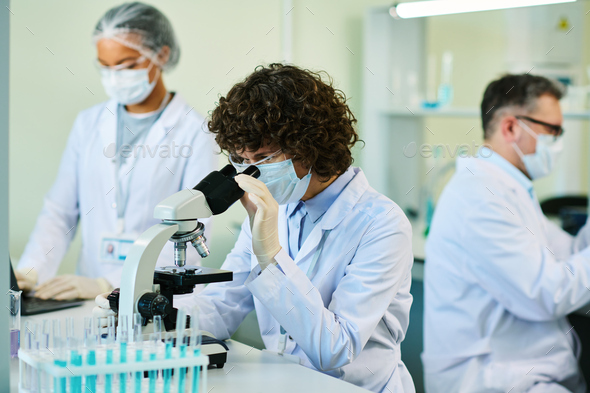 Mature brunette woman in lab coat and mask looking in microscope - Stock Photo - Images
