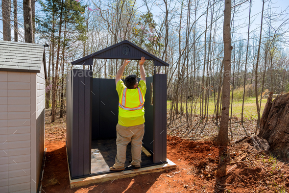 A worker is putting together a plastic vinyl storage shed for a backyard nearby