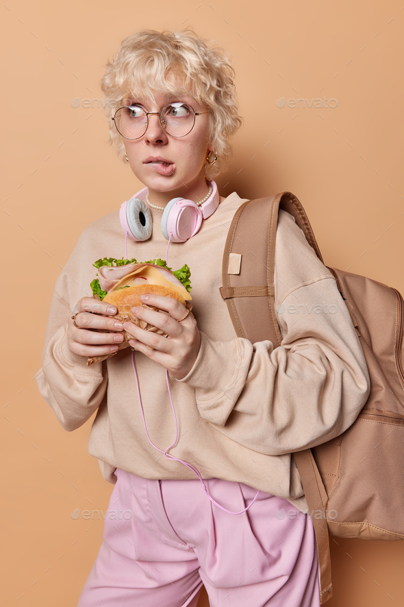 Vertical shot of hungry young female tourist eats burger bites lips stands puzzled wears pullover