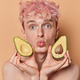 Portrait of pink haired man with freckled skin holds halves of avocado keeps lips rounded undergoes - PhotoDune Item for Sale
