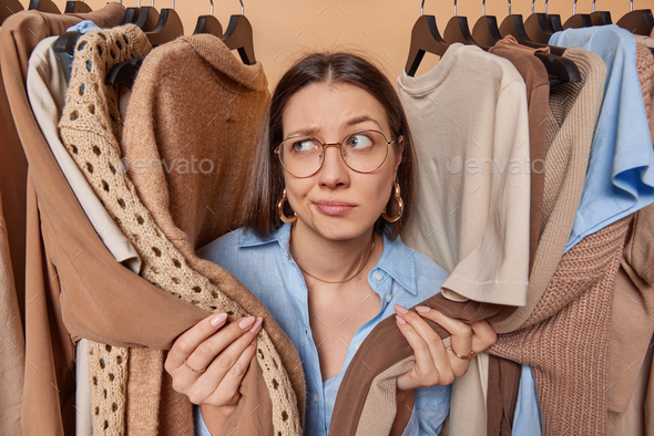 Hesitant brunette woman looks aside surrounded by garments covers herself with sleeves looks aside