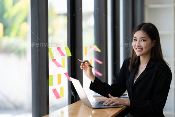 cFocused asian business woman writing idea or task on post it sticky notes on glass wall, prepare fo