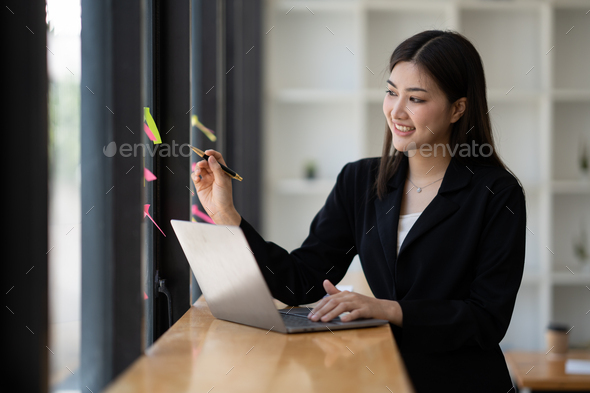 Focused asian business woman writing idea or task on post it sticky notes on glass wall, prepare for