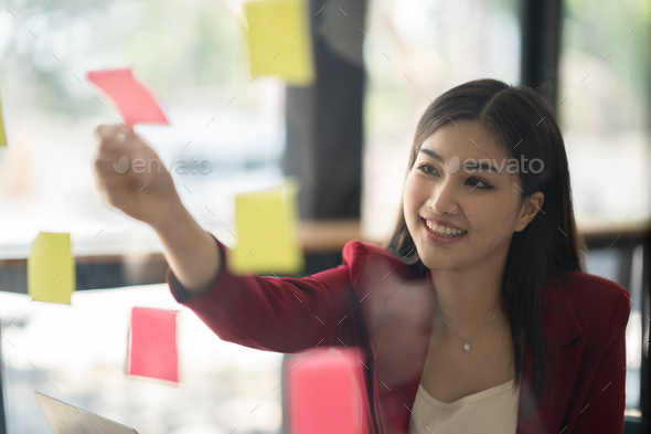 Focused asian business woman writing idea or task on post it sticky notes on glass wall, prepare for
