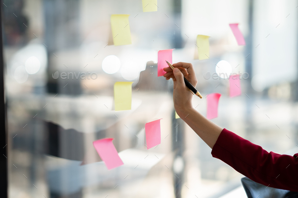 Close up view of business woman writing idea or task on post it sticky notes on glass wall, prepare