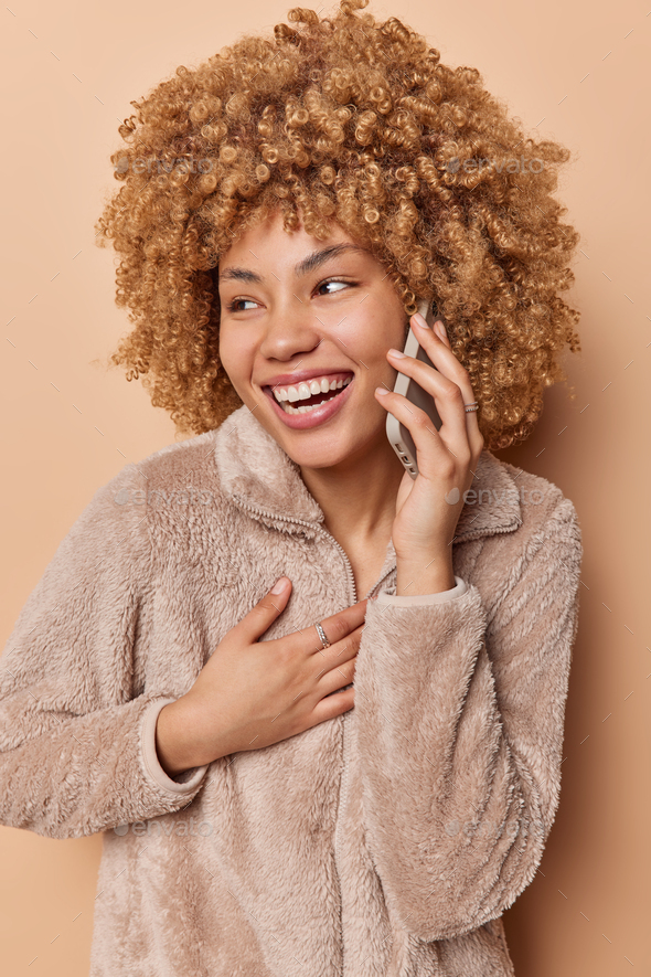 Overjoyed curly haired woman talks via smartphone and laughs positively has merry expression wears - Stock Photo - Images