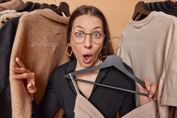 Stunned dark haired woman wears transparent eyeglasses and black dress holds clothes on hanger - Stock Photo - Images