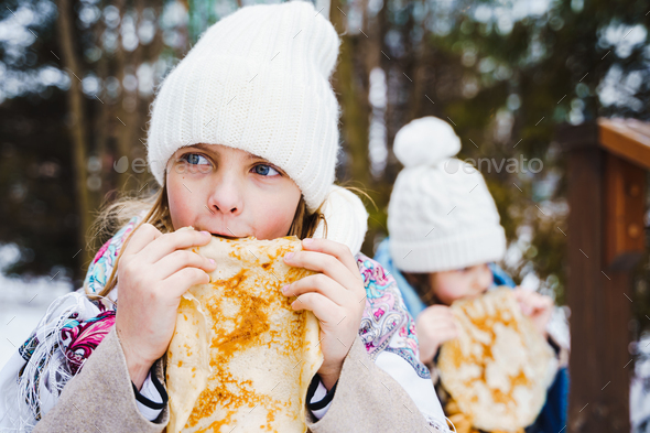 Russian national festival Maslenitsa, shrovetide. Little cute beautiful girls in headscarf eat big  - Stock Photo - Images